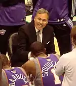 A man, wearing a black suit and a gray tie, is surrounded by basketball players as he talks to them.