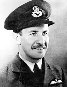 Head-and-shoulders portrait of a caucasian man in dark tie, light shirt, and dark jacket with pilot's wings on left-breast pocket, wearing peaked cap.