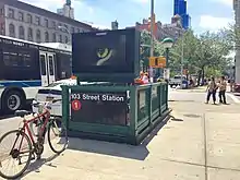 View of the rear of the exit staircase at the northwest corner of 103rd Street and Broadway. There is a sign saying "103 Street Station (1)" attached to the balustrade that surrounds the staircase. Above the sign is a video screen.