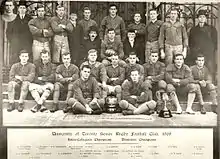 Twenty-five men, divided into three rows, pose.  The majority are wearing football uniforms, except for four executives in suits.  Two trophies are placed in front of the players.