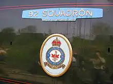 A 'Battle of Britain' class enamelled metal nameplate and crest mounted on flat metal casing covering the locomotive boiler. The nameplate forms a representation of aircraft wings, with a small rectangle attached to the middle-lower edge allowing identification as a member of the 'Battle of Briain' class. Below this is an oval-shaped plate with a crest.