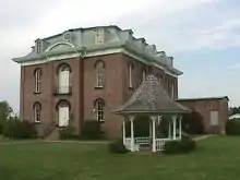 Large, ornate red brick courthouse sits on a green lawn with a gazebo in the foreground