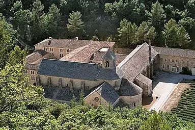 In a wooded valley is a large church with small windows and a square stone belfry. It is surrounded by ancient buildings arranged around courtyards, and a lavender garden.