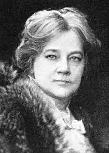 A middle-aged white woman with light hair parted center and dressed back from her face, wearing a fur wrap