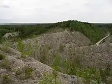 A colour photograph of the un-rehabilitated Aidu open-pit mine. The lower two-thirds of the photo shows hilly land that is mostly brownish-grey. Some small, green trees are growing within this section.