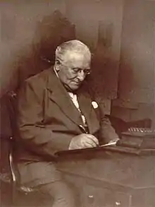 Photograph of a man, with white hair and wearing a suit, sitting working at a desk