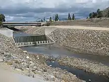 A muddy flow of water drops over an artificial concrete waterfall, from a tree-lined riverbed to a riprap-lined channel.