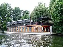 A colour image Gilmour's houseboat and studio the Astoria, anchored in a river. The background is green forest and it is a bright sunny day.