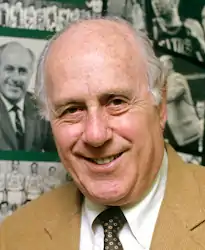 A man, wearing a brown coat, white shirt and black tie, standing in front of a collage.