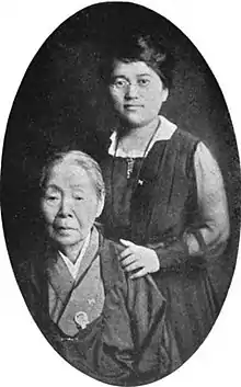 Two Japanese women in an oval portrait frame. The standing woman is younger, wearing a Western-style dress and glasses; her hands are on the shoulders of an older woman, wearing a Japanese-style dress.