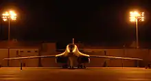 Front view of B-1 parked on ramp at night. Nearby yellow flood lights illuminate the area. In the background are buildings