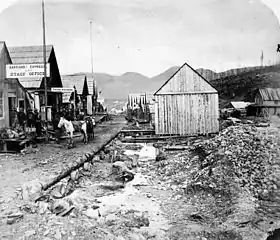 A black and white photo of a Barkerville street. On the left, a series of single-story houses follow the road into the distance; the closest one bears a sign for the "Barnard Express." In the centre, the road edges onto a shallow ditch, flanked by rocks and mining rubble. A lone one-story building with a sloped roof stands at the end of the ditch, with its windowless side wall facing the viewer. To the right of the photo, more single-story structures are seen some 50 metres away.