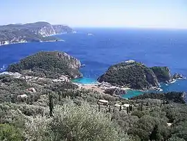 The bay of Palaiokastritsa. The rock in the sea visible near the horizon at the top centre-left of the picture is considered by the locals to be the mythical petrified ship of Odysseus. The side of the rock toward the mainland is curved in such a way as to resemble the extended sail of a trireme. The historic monastery of Palaiokastritsa can be seen at the top of the hill of the peninsula to the right of the picture.