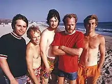 A photo of five men are standing on the beach looking at the camera. Three of the men are shirtless