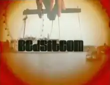 The word "Bedsitcom" written in a large, black font over a bright landscape of the south bank of the River Thames. The word is being lifted up by puppet strings held by a human hand. There is a red-orange circular glow around the image.