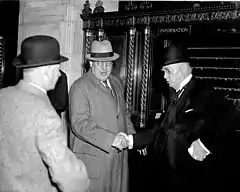 Two older white men in suits, hats, and eyeglasses shake hands.  The one on the left looks towards the foreground, where a third white man in a hat looks back, his back to the camera.