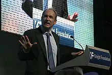 Berners-Lee speaking in front of a slide projection with right hand extended