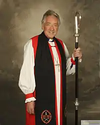 An Anglican bishop with a crosier, wearing a rochet under a red chimere and cuffs, a black tippet, and a pectoral cross