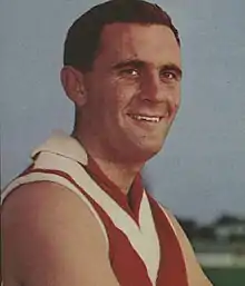 A dark haired white man smiles off camera while wearing a red woolen football jumper featuring a white chevron