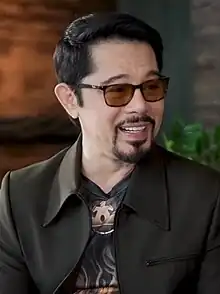 Christopher de Leon wearing a black suit jacket and a print shirt, with tinted glasses, grinning and looking left of camera