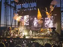 A brightly lit concert stage at sunset with puffs of smoke, television screens on the backdrop, and an audience watching.