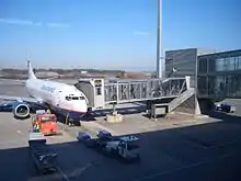 A jet passenger aircraft on the left of the image parked at a glass jetbridge that runs on the right of the image. The plane is surrounded by three aircraft handling cars