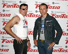 Two men in their mid-20s stand with their hands at their hips and facing the camera. The man on the left, Jason Trost, has short brown hair and wears an eyepatch, a sleeveless hooded sweatshirt, and black jeans. The man on the right, Brandon Trost, also has short brown hair, wears a denim jacket over a gray t-shirt with an unreadable graphic, and blue jeans.