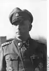 A man wearing a military uniform, peaked cap and neck order, in the shape of a cross. His cap has an emblem in shape of a human skull and crossed bones.