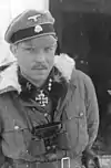 A man wearing a military uniform, coat with fur collar, peaked cap, neck order in the shape of a cross and a pair of binoculars around his neck. His cap has an emblem in shape of a human skull and crossed bones.