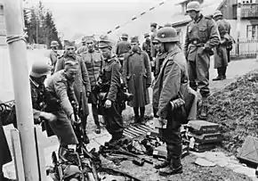 A black and white photograph of soldiers in helmets watching other soldiers laying down rifles in a pile