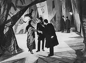 A man shakes hands with a woman, while another man looks on between them. The three figures stand in the iddle of a city street, with brick walls in twisted and distorted shapes, and shadows and streaks of light painted onto the walls and ground.