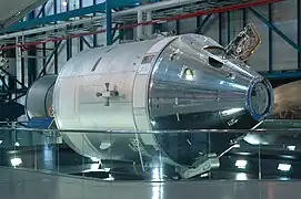 A metallic gray space capsule connected to a cylindrical module lies on its side as a museum exhibit