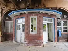 A brick station house, at street level, with an angled bay window. The windows have white trimming, while the doors and house cornice have blue trimmings.