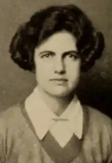 A young white woman with short dark thick hair, wearing a v-necked sweater over a collared shirt, pinned at throat