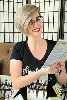 A woman with short blonde hair, red lipstick, and glasses wearing a black t-shirt, holding a book, and smiling