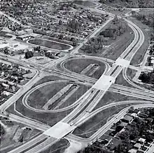 Black and white aerial photo of a half-cloverleaf highway interchange surrounded by manicured lawns and suburban development