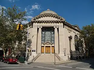 The front entrance of a hexagonal building capped by a dome is visible, facing a street-corner. The entranceway is framed by large stone columns and flanked by metal seven branched menorahs on each side. There are four wooden doors, one on each side and two in the middle, topped by a large arched stained-glass window. A stone stairway with metal railings on each side leads up from the sidewalk to the doors.