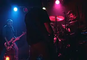 An Australian rock band, the Cosmic Psychos, performing onstage. The dark stage is lit up by coloured lights. Three performers are visible: an electric bass player, an electric guitarist, and a drummer behind a drumkit.