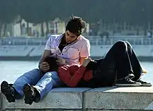 A young couple sits on a bench. The woman is lying down with her head resting on the man's lap.