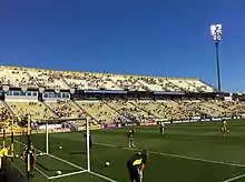 A soccer field with a multi-level stadium visible on one side, a soccer goal, and some players warming up on the field.