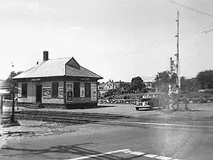 A one-story wooden train station next to a two-track rail line at a grade crossing