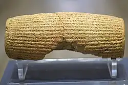 Rear view of a barrel-shaped clay cylinder resting on a stand. The cylinder is round with square edge very close up. It is covered with lines of cuneiform and has a hollow centre. The centre is lined with plate measuring around 10 millimeters in thickness.text