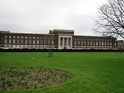 Very wide flat-roofed mulberry brick building with three rows of widows, portico is Portland stone with four white columns