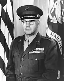 A stocky Caucasian man with brown hair and thick glasses in military uniform in front of a flag