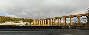 A curving stone viaduct bathed in sunshine on the right of the image. Houses and a small valley shown on the left.