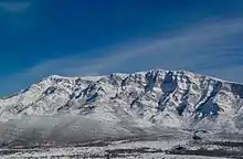 Photograph of the section of the Dinara massif 1500 meters above sea level and up, including the Samograd outcropping, with its foothills and plain, all covered in snow, and a blue sky.