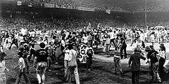 A black and white image of a crowd of young adults, mostly white men, walking around a baseball field and cheering. Some are holding signs, but what the signs say is unclear.