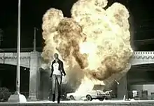 A woman is walking away from an exploding car underneath a bridge. The entire image is in black and white, with the exception of the fireball rising from the car.