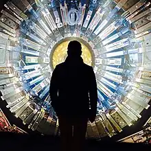 An outline of a man's shadow standing in front of a large metallic circle that is covered in machine parts