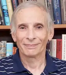 headshot of an older white man in a polo shirt in front of a bookshelf